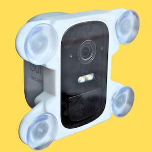 May include: A white security camera mount featuring four clear suction cups on the corners. The central black panel contains a camera lens and two LED lights. The side of the white plastic housing displays the partial text SECURITY.