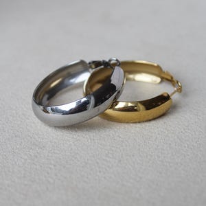 Könnte beinhalten: Zwei Creolen, eine silberne und eine goldene, werden auf einer hellen Oberfläche gezeigt. Die Ohrringe haben ein abgerundetes, dickes Design. Der silberne Ohrring befindet sich im Vordergrund und reflektiert das Licht, während sich der goldene Ohrring dahinter befindet.