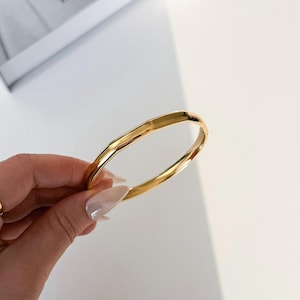 Könnte beinhalten: Ein goldfarbenes Armreif-Armband mit strukturierter Oberfläche. Das Armband wird vor einem weißen Hintergrund gehalten, um seine polierte Oberfläche und sein schlichtes Design hervorzuheben. Das Schmuckstück ist ein klassisches Accessoire.