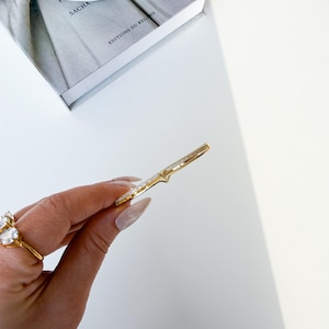 May include: A gold ring with a V-shaped design, set with small, clear gemstones. The ring is held in a hand with long, manicured nails. Another ring with a large, clear gemstone is also visible.
