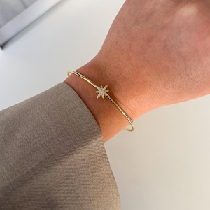 May include: A gold-coloured bracelet featuring a starburst design. The thin, delicate bracelet has a small starburst charm at its centre, encrusted with tiny, clear crystals. The bracelet is worn on a wrist, with a neutral-toned sleeve visible in the background.