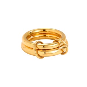 Könnte beinhalten: Ein goldfarbener Ring mit einem einzigartigen Design. Der Ring besteht aus drei übereinanderliegenden Bändern, die durch ein dekoratives Element verbunden sind. Die Bänder haben eine glatte, polierte Oberfläche, die das Licht reflektiert. Der Ring ist ein Statement-Schmuckstück.