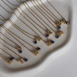 Könnte beinhalten: Eine Sammlung von goldenen Halsketten mit Buchstabenanhängern und blauen bösen Augenperlen. Die Halsketten werden auf einer weißen Oberfläche präsentiert.