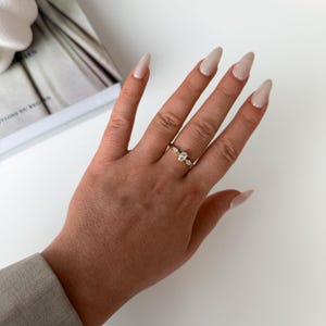 Könnte beinhalten: Ein goldener Ring mit einem ovalförmigen Mittelstein und kleineren Akzentsteinen. Der Ring befindet sich an einer Hand mit langen, polierten Nägeln. Der Hintergrund umfasst ein Buch und eine weiße Oberfläche.
