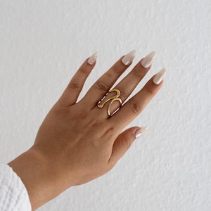Könnte beinhalten: Ein goldfarbener Ring mit abstraktem Design wird an einer Hand mit langen, polierten Nägeln getragen. Der Ring hat eine fließende, offene Form, und die Hand ist vor einem weißen Hintergrund positioniert.