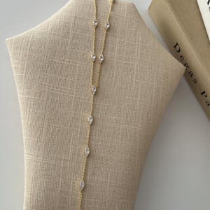 Könnte beinhalten: Eine goldfarbene Halskette mit klaren, oval geformten Edelsteinen. Die Halskette wird auf einer beigefarbenen Schmuckbüste präsentiert. Die Edelsteine sind gleichmäßig entlang der Kette angeordnet und bilden ein zartes Design. Im Hintergrund sind Bücher zu sehen.