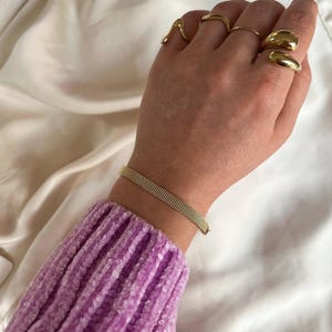 Könnte beinhalten: Ein goldenes Kettenarmband mit einem zarten, gewebten Design. Das Armband befindet sich am Handgelenk einer Person, die einen lila Pullover trägt.