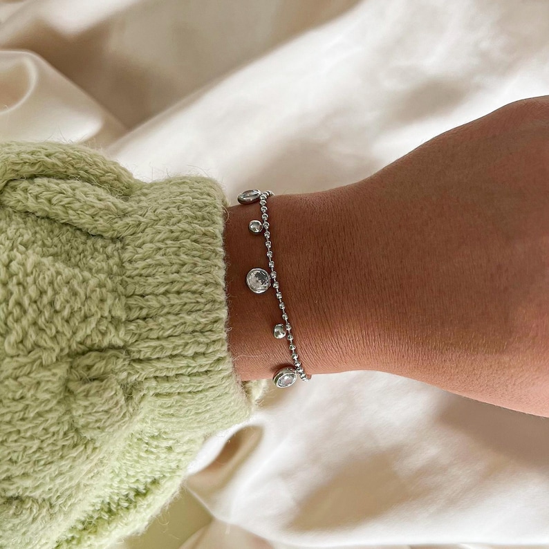 May include: A delicate silver bracelet with several clear, round, and teardrop-shaped gemstones. The bracelet is on a wrist, next to a light green knitted sleeve. The background is a white, silky fabric.