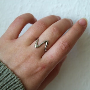 Peut inclure: Une bague argentée avec un motif ondulé. La bague est portée au doigt, le motif ondule de haut en bas. La bague est en métal brillant et a une surface lisse. C'est un bijou.