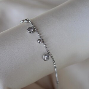 May include: A delicate silver bracelet with a beaded chain design. The bracelet features three clear, round, bezel-set gemstones and small silver beads. The jewelry is displayed on a soft, off-white fabric.