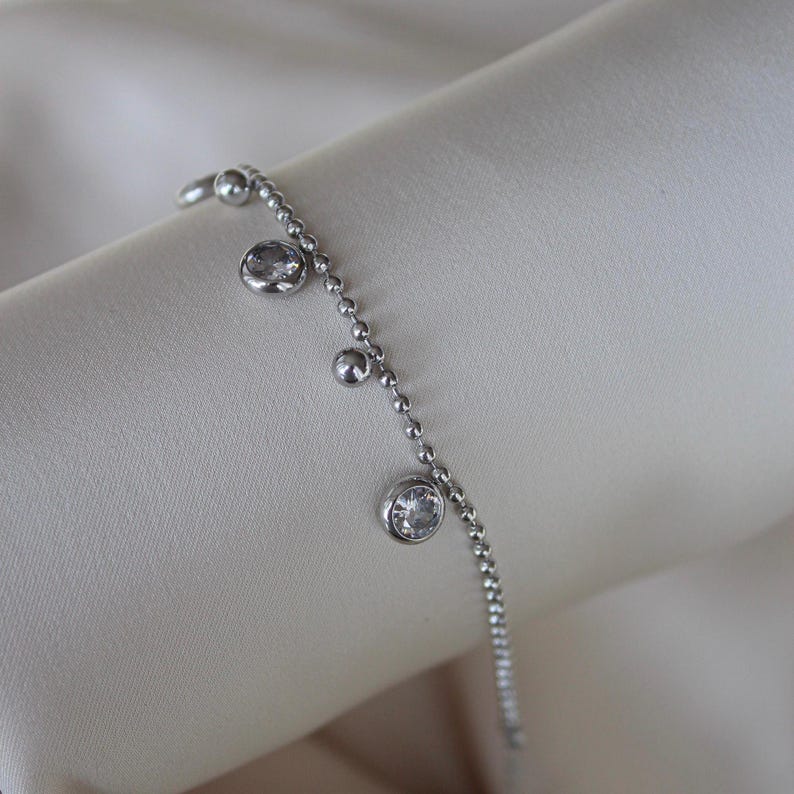 May include: A delicate silver bracelet with a beaded chain design. The bracelet features three clear, round, bezel-set gemstones and small silver beads. The jewelry is displayed on a soft, off-white fabric.