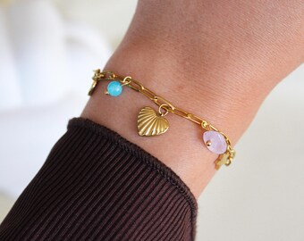 Könnte beinhalten: Goldfarbenes Armband mit Anhängern, darunter ein herzförmiger Anhänger, eine hellblaue Perle und ein rosa Edelstein. Das Armband wird am Handgelenk getragen, mit einem braunen Ärmel im Vordergrund. Das Armband hat ein zartes, feminines Design.