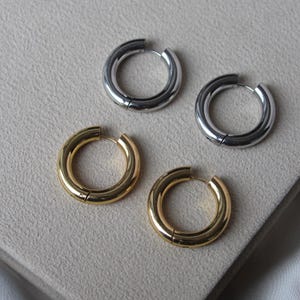 Könnte beinhalten: Vier Creolen, zwei silberne und zwei goldene. Die Ohrringe sind dick und kreisförmig, mit einem Klappverschluss. Die goldenen Ohrringe haben ein glänzendes Finish, während die silbernen Ohrringe poliert aussehen. Die Ohrringe liegen auf einer hellgrauen Oberfläche.