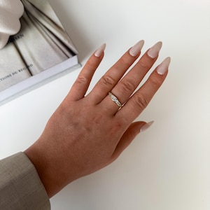 Könnte beinhalten: Ein goldener Ring mit drei klaren Steinen. Der Ring befindet sich an einer Hand mit langen, polierten Nägeln. Im Hintergrund ist ein Buch mit dem Titel "CTIONS DU REG" zu sehen.