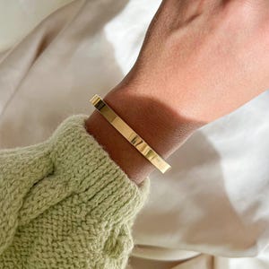 Könnte beinhalten: Ein goldfarbenes Armreif-Armband mit einer glatten, flachen Oberfläche. Das Armband wird am Handgelenk getragen.