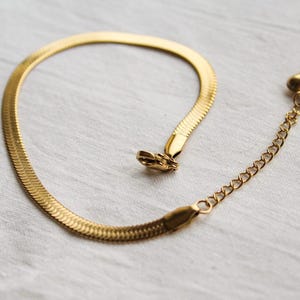 Könnte beinhalten: Ein goldfarbenes Fischgräten-Kettenarmband mit flachem, flexiblem Glieddesign. Das Armband verfügt über einen Karabinerverschluss und eine verstellbare Verlängerungskette mit kreisförmigen Gliedern für einen sicheren und anpassbaren Sitz.