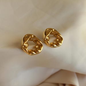Könnte beinhalten: Ein Paar goldfarbene Ohrringe mit einer abstrakten, organischen Form. Die Ohrringe haben ein großes, offenes Kreisdesign.