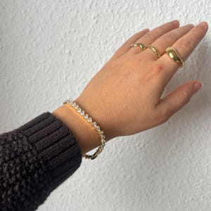 Könnte beinhalten: Ein goldfarbenes Armband mit klaren, tropfenförmigen Steinen wird am Handgelenk getragen. Die Hand trägt auch drei goldene Ringe. Die Person trägt einen dunkelgrauen Pullover.