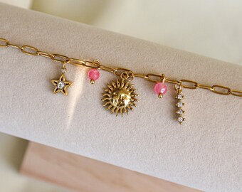 Könnte beinhalten: Goldfarbenes Kettenarmband mit mehreren Anhängern. Zu den Anhängern gehören ein Stern mit einem klaren Stein, eine Sonne, rosa Perlen und ein vertikaler Anhänger mit klaren Steinen. Das Armband wird auf einer neutralen Oberfläche präsentiert.