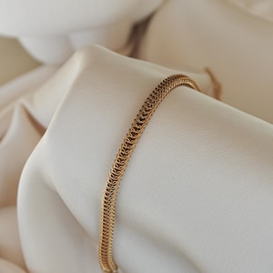 Könnte beinhalten: Ein goldfarbenes Kettenarmband mit einem gewebten Design. Das Armband wird auf weißem Stoff präsentiert, wodurch seine komplizierten Details und sein warmer metallischer Ton hervorgehoben werden. Das Schmuckstück ist ein schlichtes, aber elegantes Accessoire. Armbandlänge ca. 20 cm.