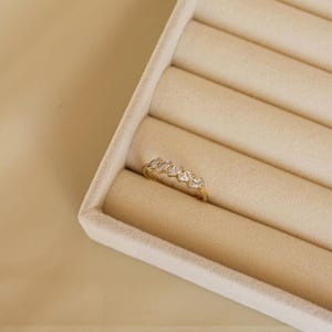 Könnte beinhalten: Ein goldener Ring mit fünf klaren, oval geformten Edelsteinen. Der Ring wird in einer beigen Schmuckschatulle mit weicher, samtiger Textur präsentiert. Das Design ist zart und elegant.