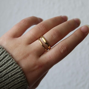 Könnte beinhalten: Zwei goldene Ringe werden an einem Finger getragen. Die Ringe sind schlichte Bänder mit poliertem Finish. Die Ringe bestehen aus einem glänzenden, gelben Metall. Der Hintergrund ist eine neutrale, cremefarbene Farbe.