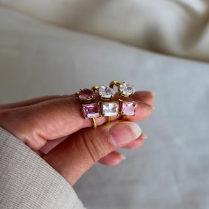 Könnte beinhalten: Eine Sammlung goldfarbener Ringe, jeder mit einem farbigen Edelstein. Die Edelsteine sind in Rosa-, Weiß- und Lilatönen gehalten und haben verschiedene Schliffarten. Die Ringe werden vor einem weichen, neutralen Hintergrund präsentiert.