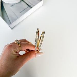 May include: A gold ring with two clear stones and two gold bangles with clear stones are held against a white background. The bangles and ring are part of a jewelry collection.