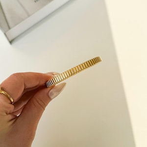 Può includere: Un braccialetto color oro con un design a coste. Il braccialetto è tenuto da una persona con un anello d'oro al dito. Lo sfondo è bianco.