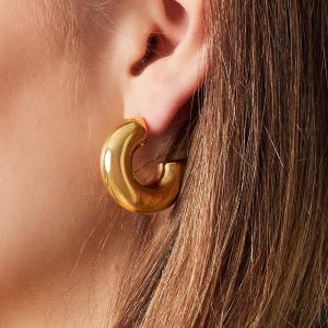 Könnte beinhalten: Goldene Creolen mit einem dicken, abgerundeten Design. Die Ohrringe werden am Ohr einer Person getragen.