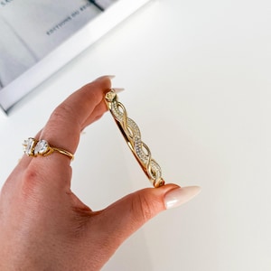 Könnte beinhalten: Goldfarbenes Armreif-Armband mit geflochtenem Design, verziert mit klaren Kristallen. Ein goldener Ring mit drei klaren Steinen ist ebenfalls zu sehen. Der Schmuck wird auf einer weißen Oberfläche präsentiert.