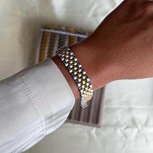 Könnte beinhalten: Ein zweifarbiges Armband mit abwechselnd goldenen und silbernen Gliedern. Das Armband im Uhrenarmbandstil befindet sich an einem Handgelenk, mit einem weißen Hemdärmel im Hintergrund. Das Armband ist aus Metall und hat einen Verschluss.