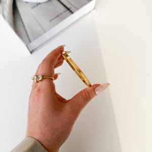 Könnte beinhalten: Ein goldfarbenes, quadratisch geformtes Armreif-Armband wird in einer Hand gehalten, zusammen mit einem goldenen Ring mit drei klaren Steinen. Der Schmuck befindet sich vor einem weißen Hintergrund, mit einem Buch in der oberen linken Ecke.