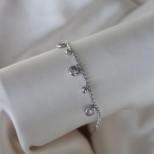 May include: A delicate silver bracelet with a series of small, round beads and several dangling clear, round gemstones. The bracelet is displayed on a soft, off-white fabric, highlighting its elegant design. A simple and refined piece of jewelry.