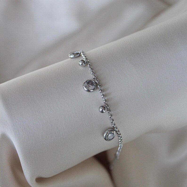 May include: A delicate silver bracelet with a series of small, round beads and several dangling clear, round gemstones. The bracelet is displayed on a soft, off-white fabric, highlighting its elegant design. A simple and refined piece of jewelry.