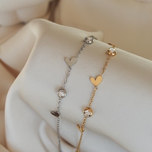 Könnte beinhalten: Zwei zarte Armbänder, eines silberfarben und eines goldfarben, jeweils mit herzförmigen Anhängern und funkelnden runden Kristallen. Die Armbänder werden auf einem weichen, weißen Stoff präsentiert, der ihr elegantes Design hervorhebt.