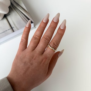 Könnte beinhalten: Ein goldfarbener Ring mit einem wellenförmigen Design, verziert mit kleinen, klaren Edelsteinen. Der Ring befindet sich an einer Hand mit langen, polierten Nägeln. Die Nägel sind in einer hellen, neutralen Farbe lackiert. Der Hintergrund ist weiß.