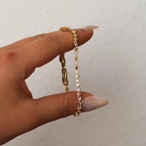 Könnte beinhalten: Ein goldfarbenes Kettenarmband mit einer Reihe sich wiederholender dreieckiger Glieder, die jeweils mit einem kleinen, klaren, funkelnden Stein besetzt sind. Das Armband hat ein filigranes, gleichmäßiges Design mit polierter Oberfläche und Verschluss.