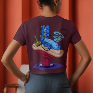 以下が含まれることがあります： アリス・イン・ワンダーランドの青いイモムシが水タバコを吸ってキノコの上に横たわっているグラフィックがプリントされた、マルーンのクロップトップです。