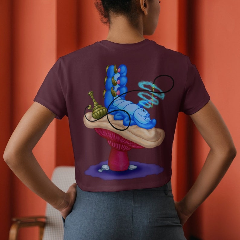 以下が含まれることがあります： アリス・イン・ワンダーランドの青いイモムシが水タバコを吸ってキノコの上に横たわっているグラフィックがプリントされた、マルーンのクロップトップです。