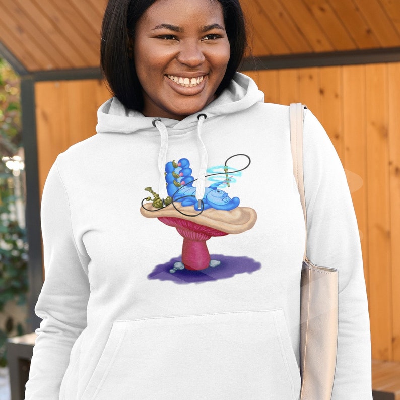 以下が含まれることがあります： 白いフード付きスウェットシャツに、不思議の国のアリスの青いイモムシがキノコの上で水タバコを吸っているグラフィックがプリントされています。