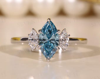 May include: A silver ring featuring a large, marquise-cut, light blue gemstone flanked by two smaller, white, marquise-cut stones. The ring is set on a white surface with a blurred background.