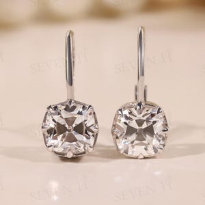 May include: A pair of silver-toned drop earrings. Each earring features a square, clear crystal with multiple facets, reflecting light. The earrings have a simple, elegant design, suitable for formal or everyday wear. The background is a soft, neutral color.