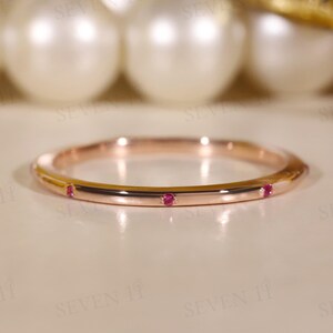 Peut inclure: Une bague en or rose avec trois petites pierres précieuses rouges rondes, espacées sur le pourtour. La bague est très polie et reflète la lumière, sur un fond flou de perles et de tissu doré.