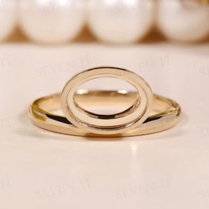 Puede incluir: Un anillo de oro pulido que presenta un engaste ovalado prominente orientado verticalmente en el centro. La banda es lisa y circular, conectándose a la base del marco ovalado. El metal tiene un acabado de oro amarillo cálido y brillante.