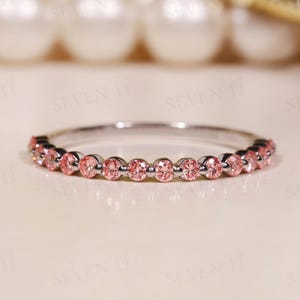Peut inclure: Une fine bague en métal argenté ornée d'une rangée continue de petites pierres précieuses rondes rose clair, serties dans une monture à griffes partagées. L'anneau est fin et poli, présentant une série de pierres roses facettées uniformes.
