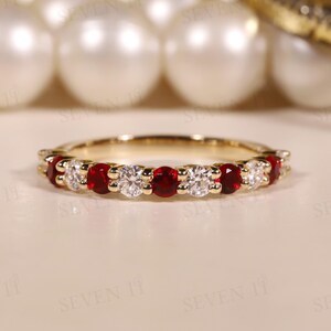 May include: A gold band ring with alternating red and clear gemstones. The ring showcases a series of small, vibrant red stones and sparkling clear stones, forming a symmetrical pattern. The ring is presented on a neutral surface.