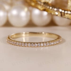 Op de afbeelding: Een gouden bandring met kleine, heldere edelstenen rondom de hele omtrek. De ring ligt op een lichtgekleurd oppervlak, met vage parels en gouden accenten op de achtergrond.