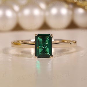 Puede incluir: Un anillo de oro con una piedra preciosa verde de talla esmeralda rectangular. El anillo tiene una fina banda de oro pulido. La piedra preciosa es el punto focal, reflejando la luz y engastada en un engaste de cuatro garras. El anillo se exhibe sobre una superficie reflectante.