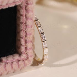 May include: A gold band ring featuring a row of clear, rectangular gemstones. The ring is presented on a pink, textured surface. The brand name "SEVEN 11" is visible in the bottom left corner.